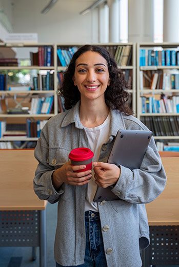 Mujer sonriente sostiene un café y una tablet frente a estanterías de libros.